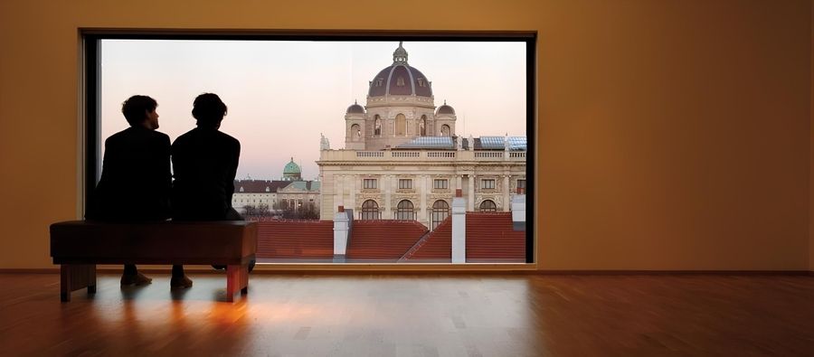 Zwei Personen in Silhouette sitzen auf einer Bank und blicken aus einem Fenster auf ein großes Gebäude.