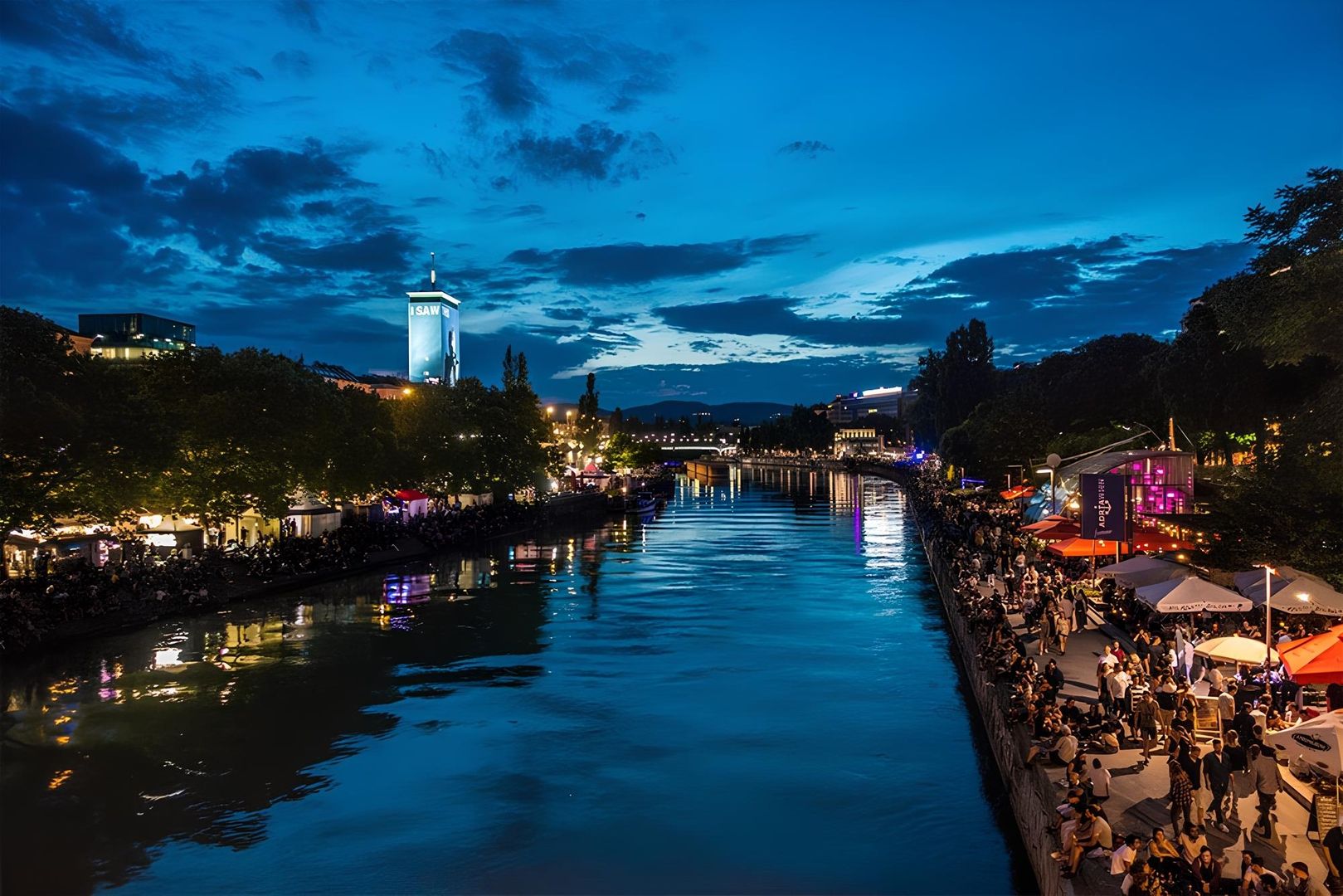 Abendlicher Blick auf den Wiener Donaukanal mit Menschen am Ufer und Lichtern die sich im Wasser spiegeln