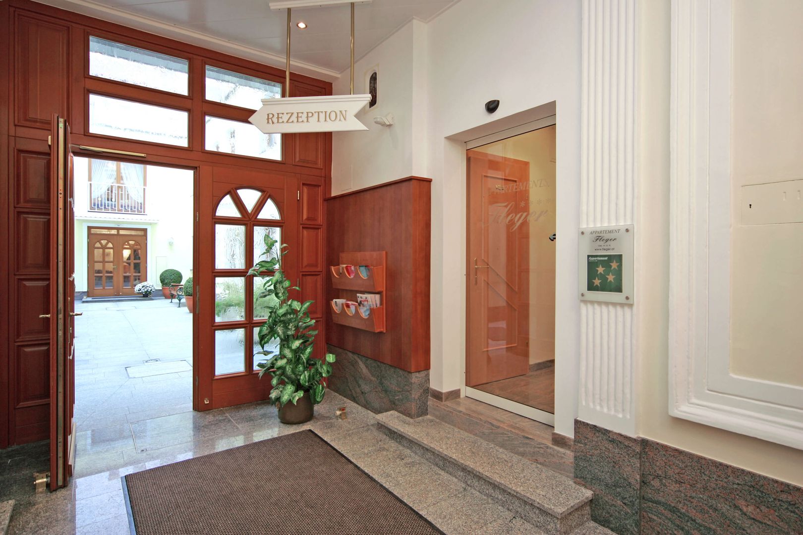 Bright hotel entrance with glass door, reception sign, brochure stand and view into the courtyard.