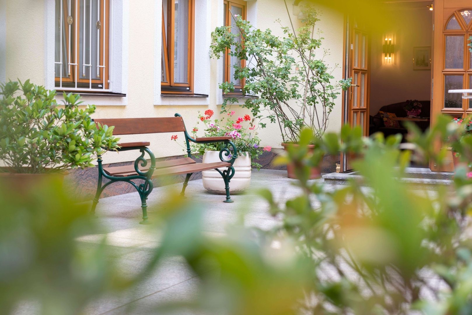 Ruhiger Innenhof mit Holzbank, blühenden Pflanzen in Töpfen und offenstehender Tür zu einem Wohnraum.