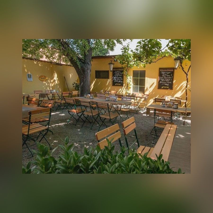 Leerer Biergarten im Schatten eines Baumes mit Holztischen und Stühlen einer gelben Hauswand und grünen Pflanzen im Vordergrund