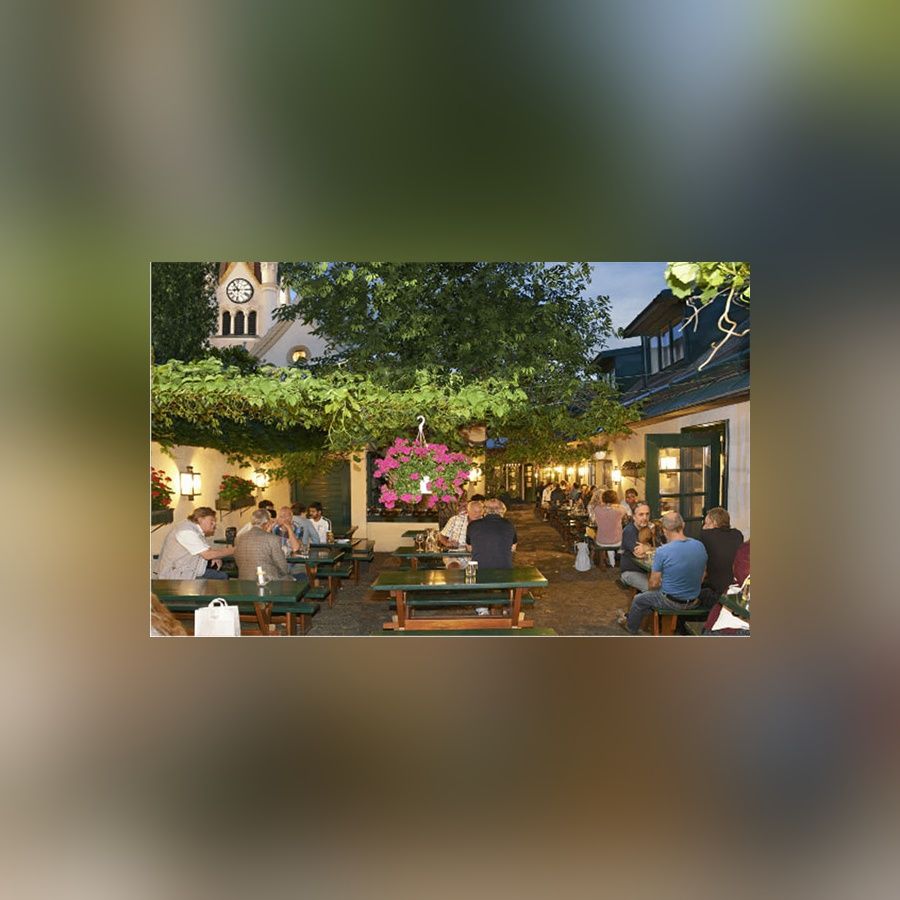Gemütlicher Biergarten am Abend mit beleuchteten Tischen und Gästen unter einem bewachsenen Vordach im Hintergrund ein Uhrturm