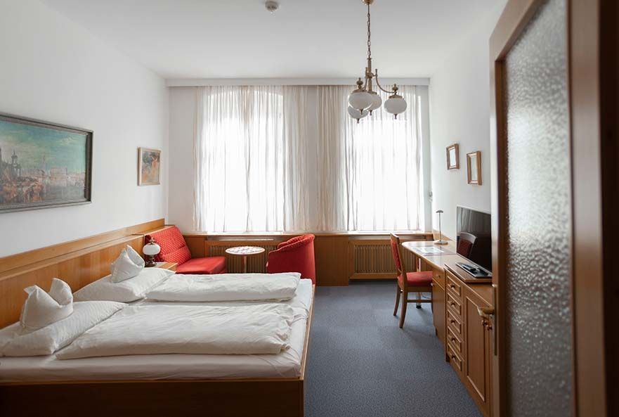 Bright hotel room with double bed, red sofa, desk and large windows with curtains.