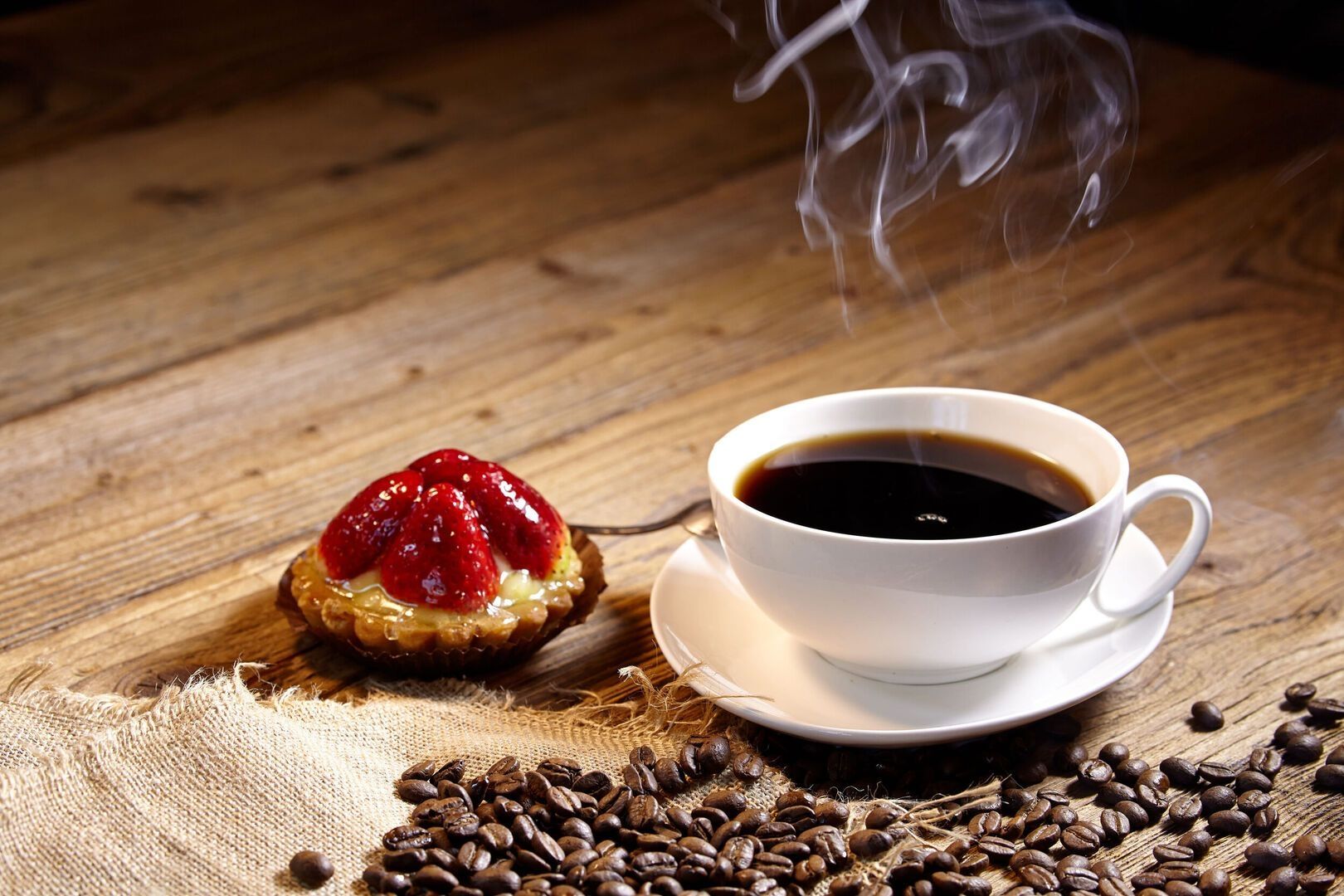 Tasse schwarzer Kaffee mit aufsteigendem Dampf neben einem Erdbeertörtchen und Kaffeebohnen auf Holztisch.