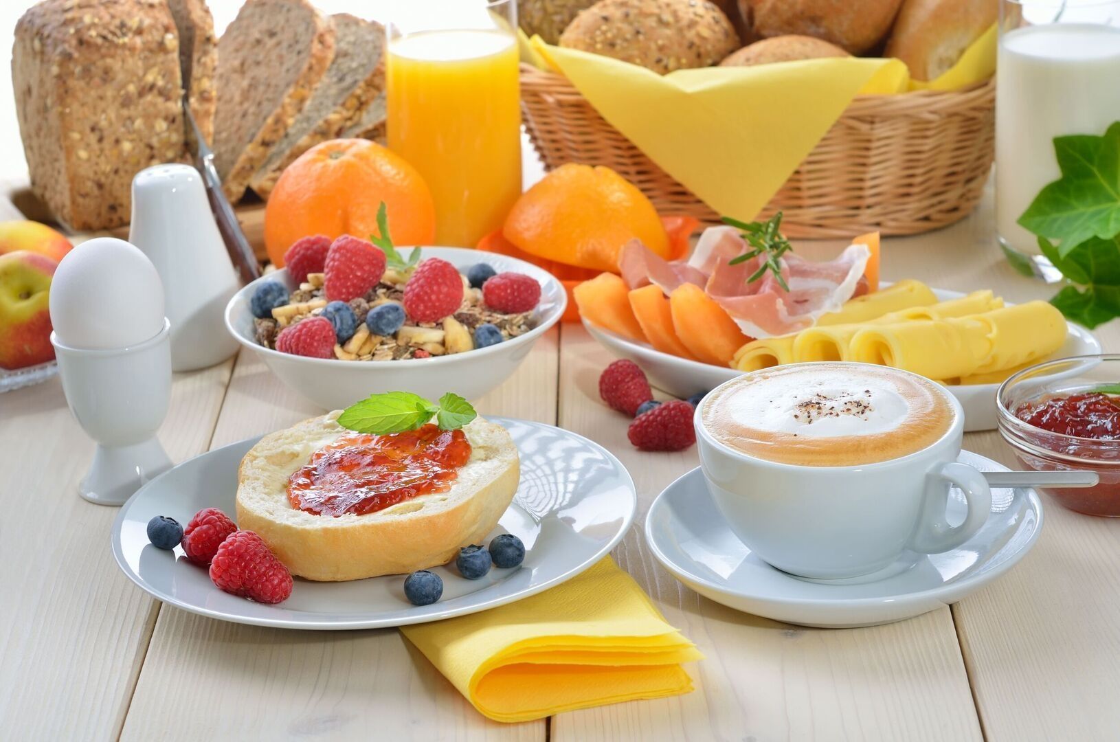 Frühstückstisch mit Brötchen, Marmelade, frischem Obst, Müsli, Käse, Schinken, gekochtem Ei, Cappuccino, Orangensaft und Brotkorb.