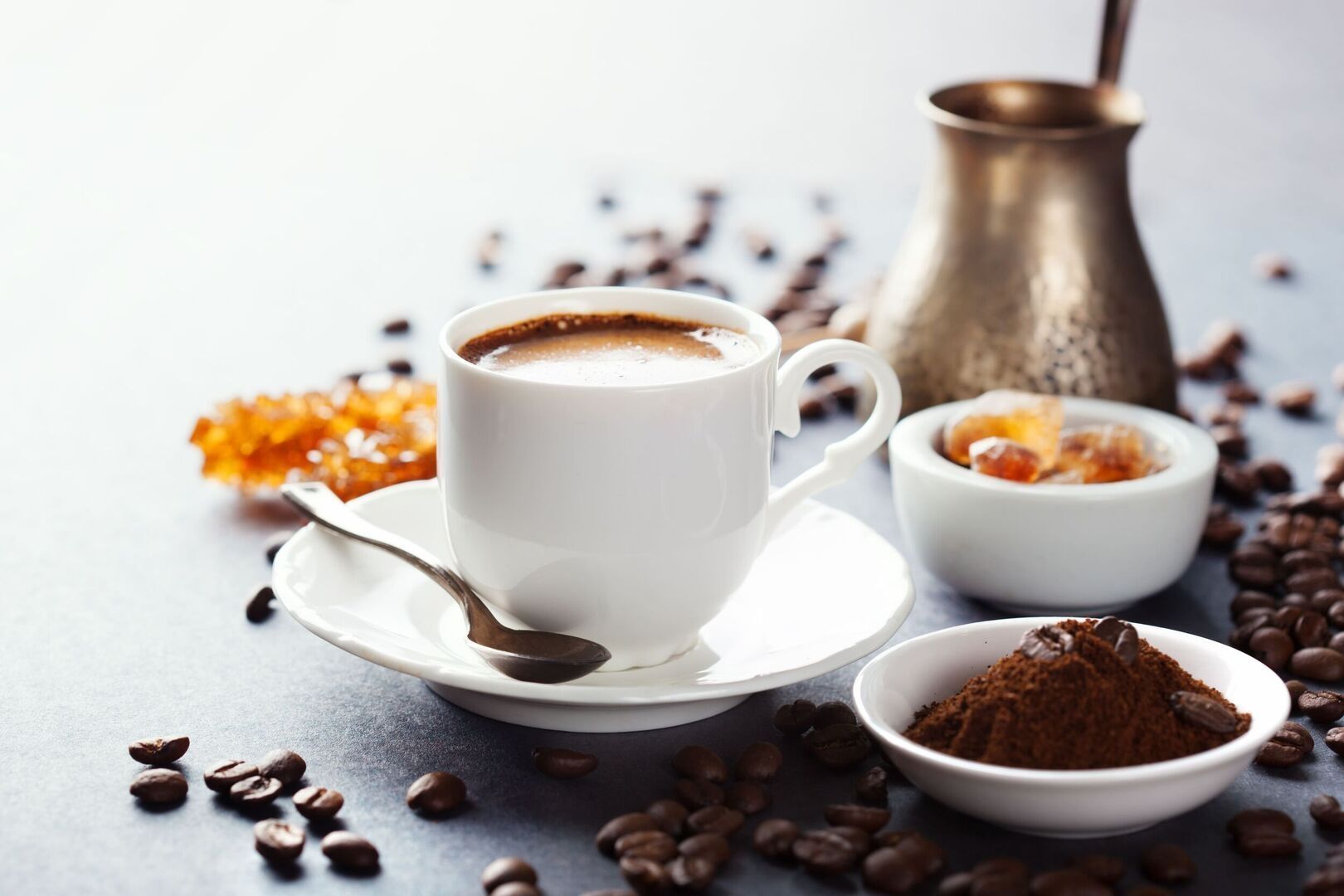 Tasse frisch gebrühter Kaffee auf Untertasse mit Löffel, daneben Kaffeepulver, Kaffeebohnen, Zucker und eine Kupferkanne.