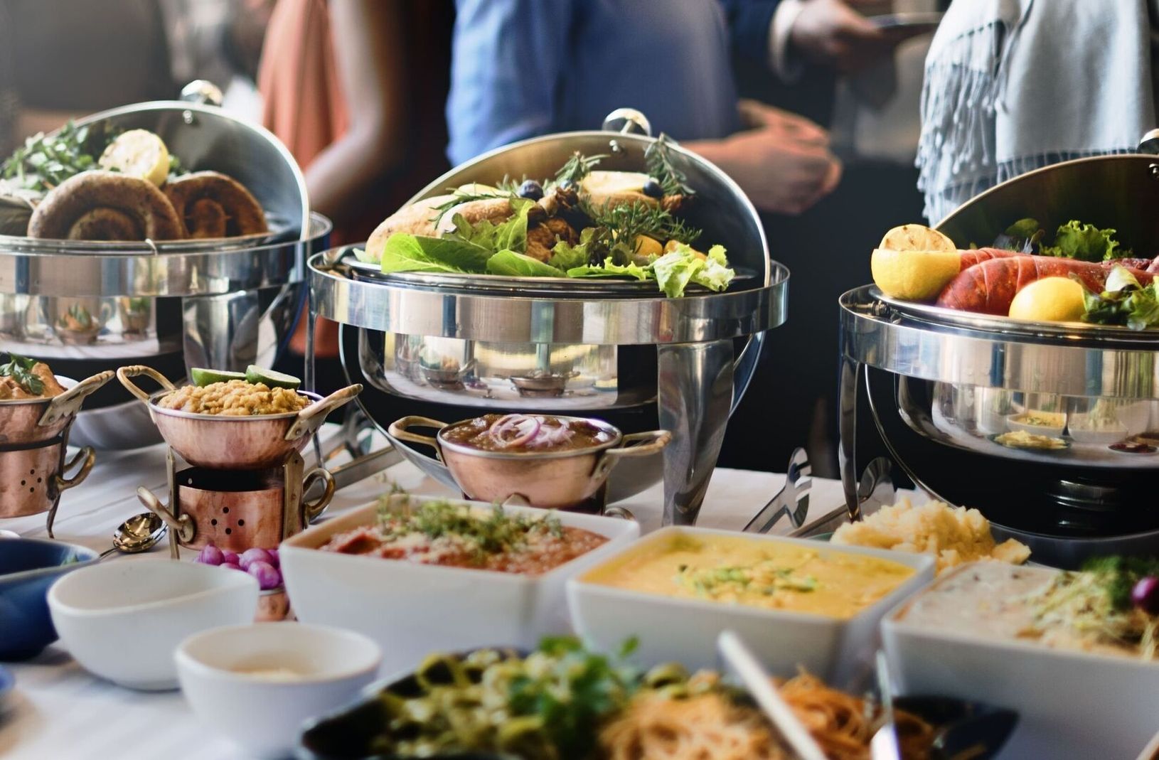 Buffettisch mit verschiedenen warmen Speisen in Metallbehältern, Saucen, Beilagen und Salaten.