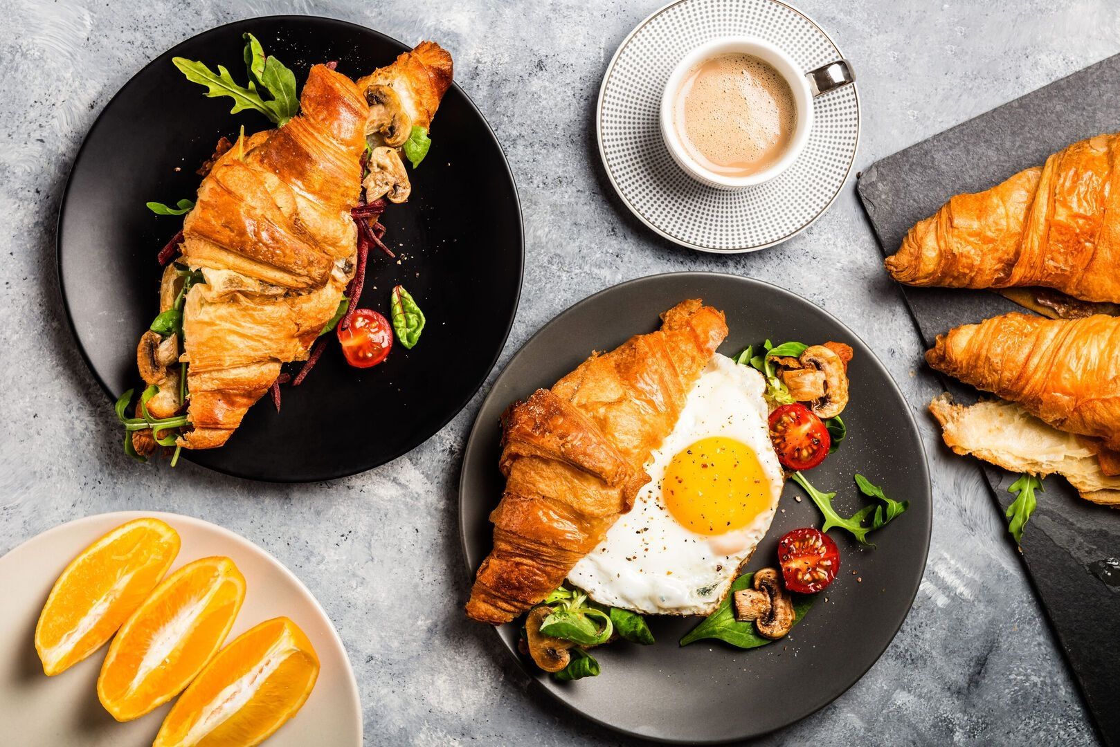 Frühstückstisch mit zwei Croissants gefüllt mit Spiegelei Pilzen Rucola und Kirschtomaten daneben eine Tasse Kaffee und Orangenscheiben