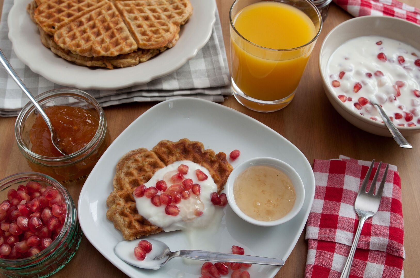 Frühstücksteller mit Waffeln, Joghurt und Granatapfelkernen, dazu Marmelade, Honig, Orangensaft und Schüssel mit Joghurt.