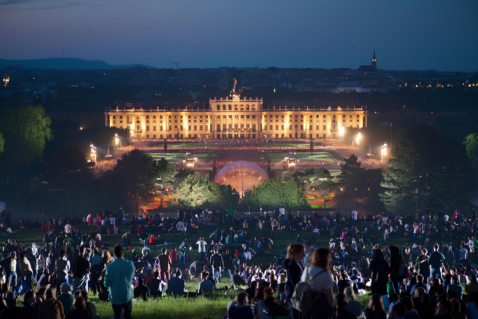 Große Menschenmenge bei einem Open Air Konzert vor dem beleuchteten Schloss Schönbrunn in der Abenddämmerung.