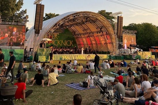 Menschen sitzen auf einer Wiese und schauen einer Band auf einer Open Air Bühne zu.