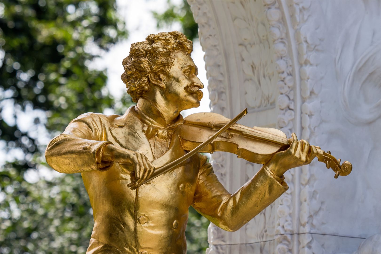 Golden statue of a violinist in front of an ornate white stone relief outdoors.